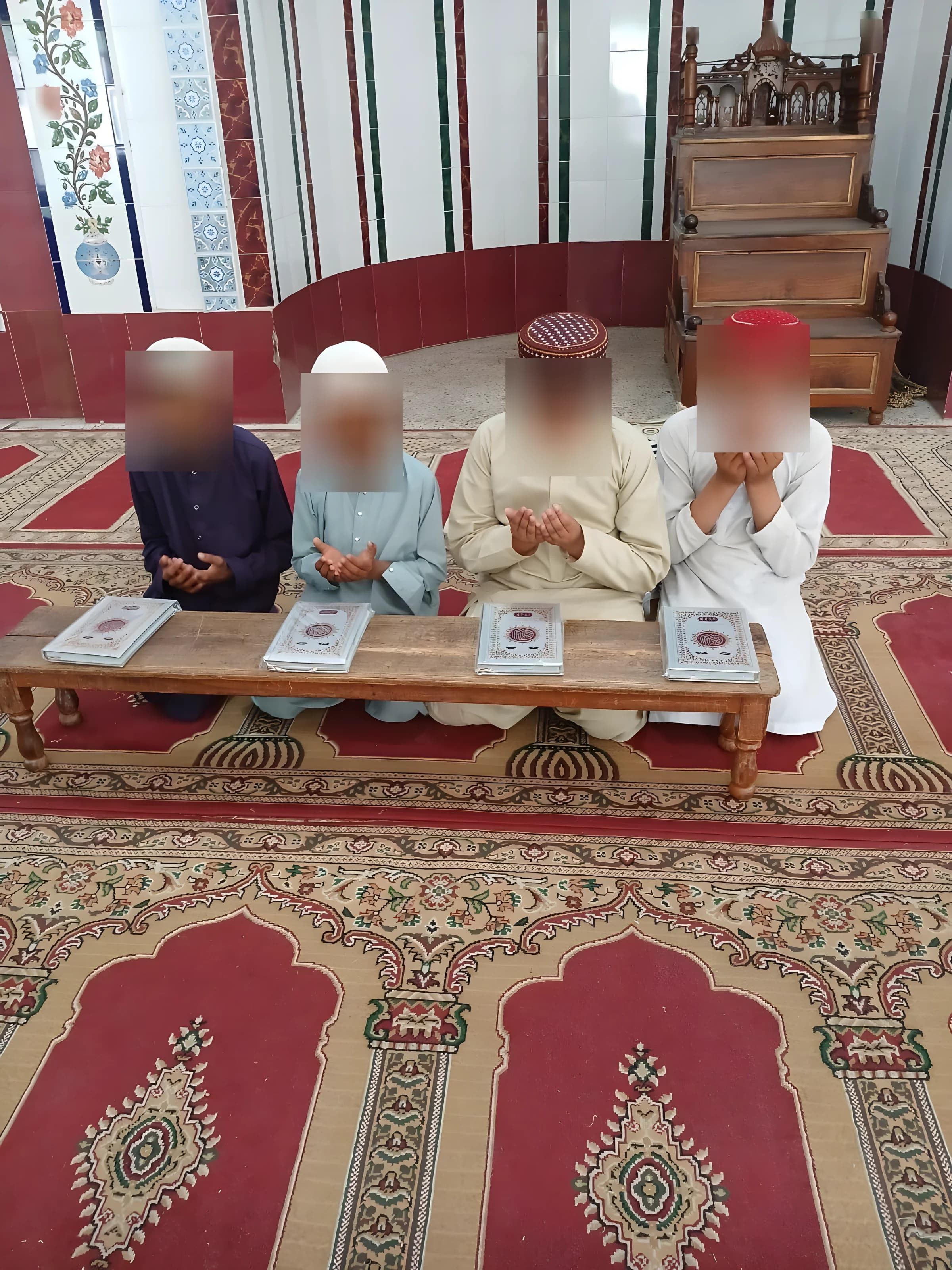 Fire gutter sitter med hendene løftet i dua bak en lav trebenk med fire Quraner