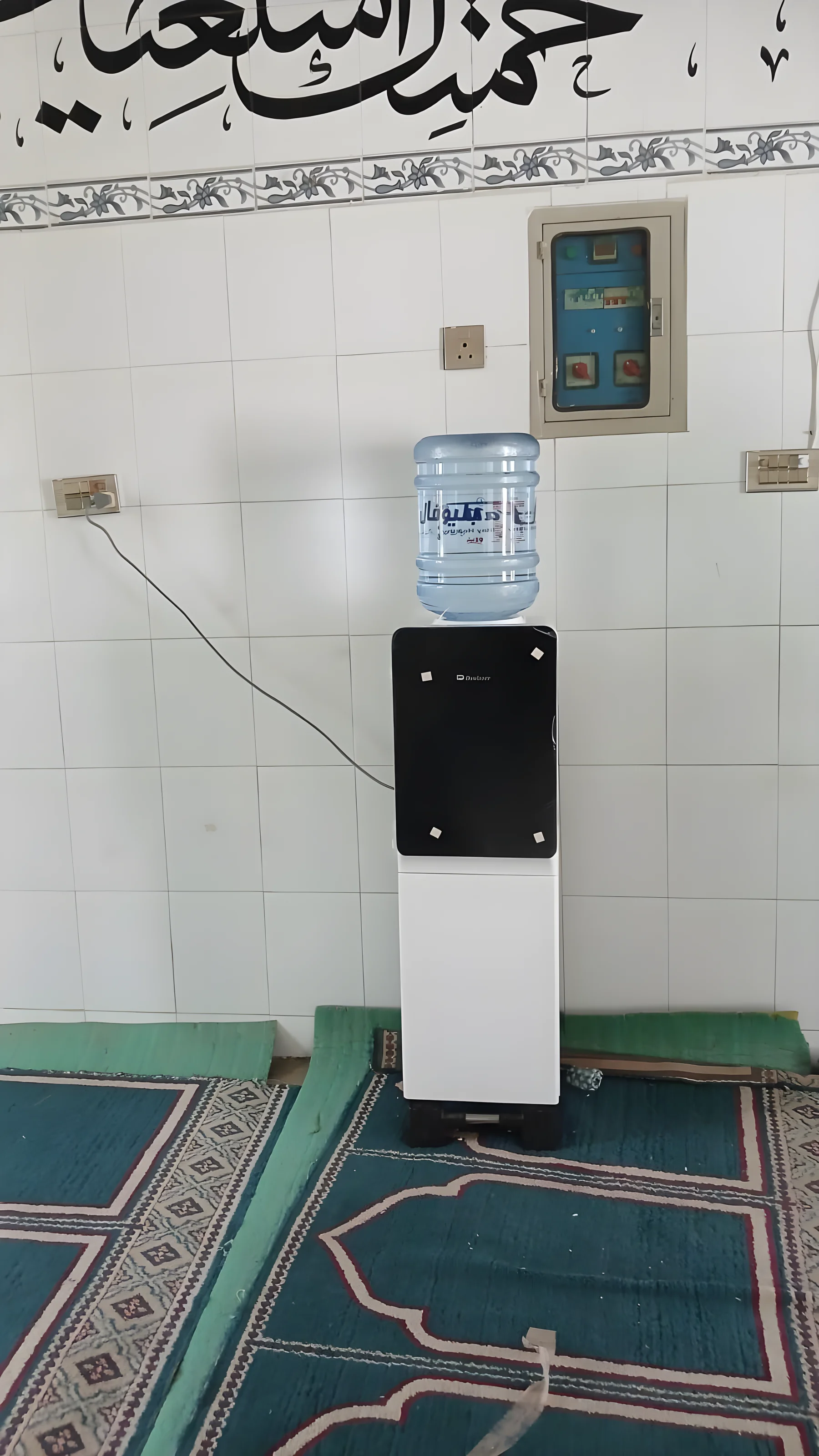 Vanndispenser med full flaske står mot en flislagt vegg i en masjid, på grønne bønnetepper